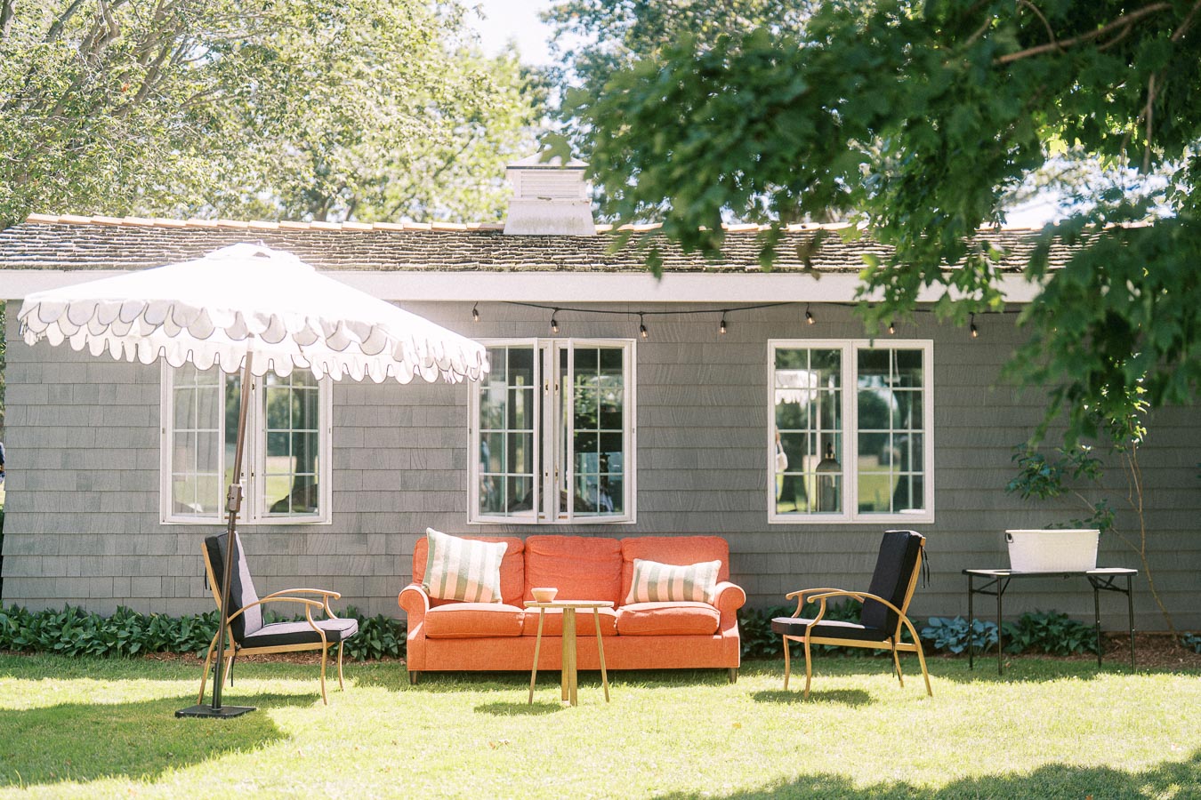 Outdoor wedding cocktail hour lounge with orange sofa, chairs, umbrella, and string lights on a private estate lawn near Chatham-Kent, Ontario.