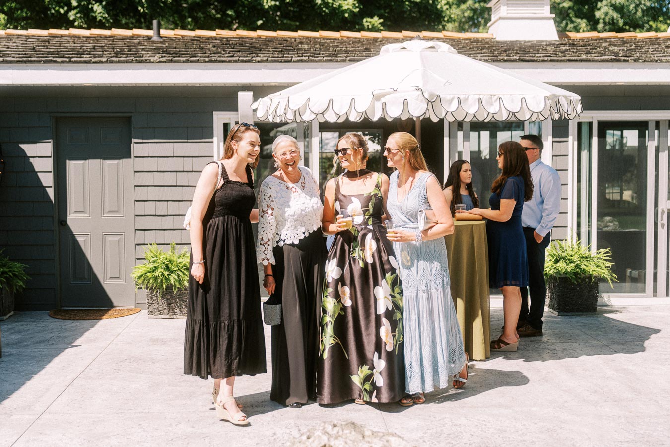 Wedding guests in formal dresses gather with drinks under a white patio umbrella during cocktail hour at a private estate near Chatham-Kent, Ontario.