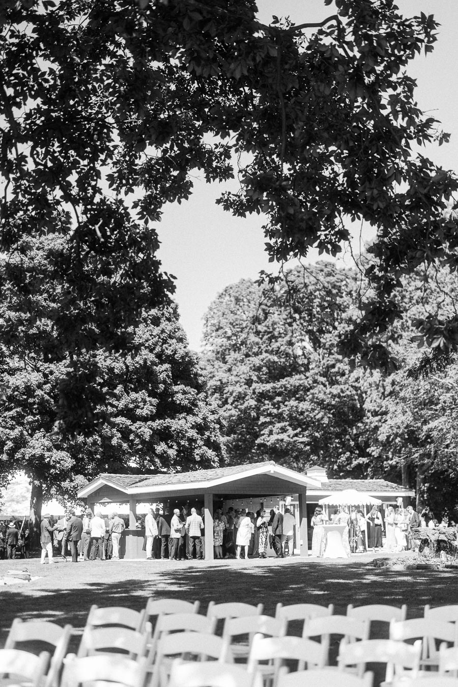 Black and white photo of wedding guests mingling during cocktail hour near a pavilion with ceremony chairs in foreground at a private estate in Ontario.