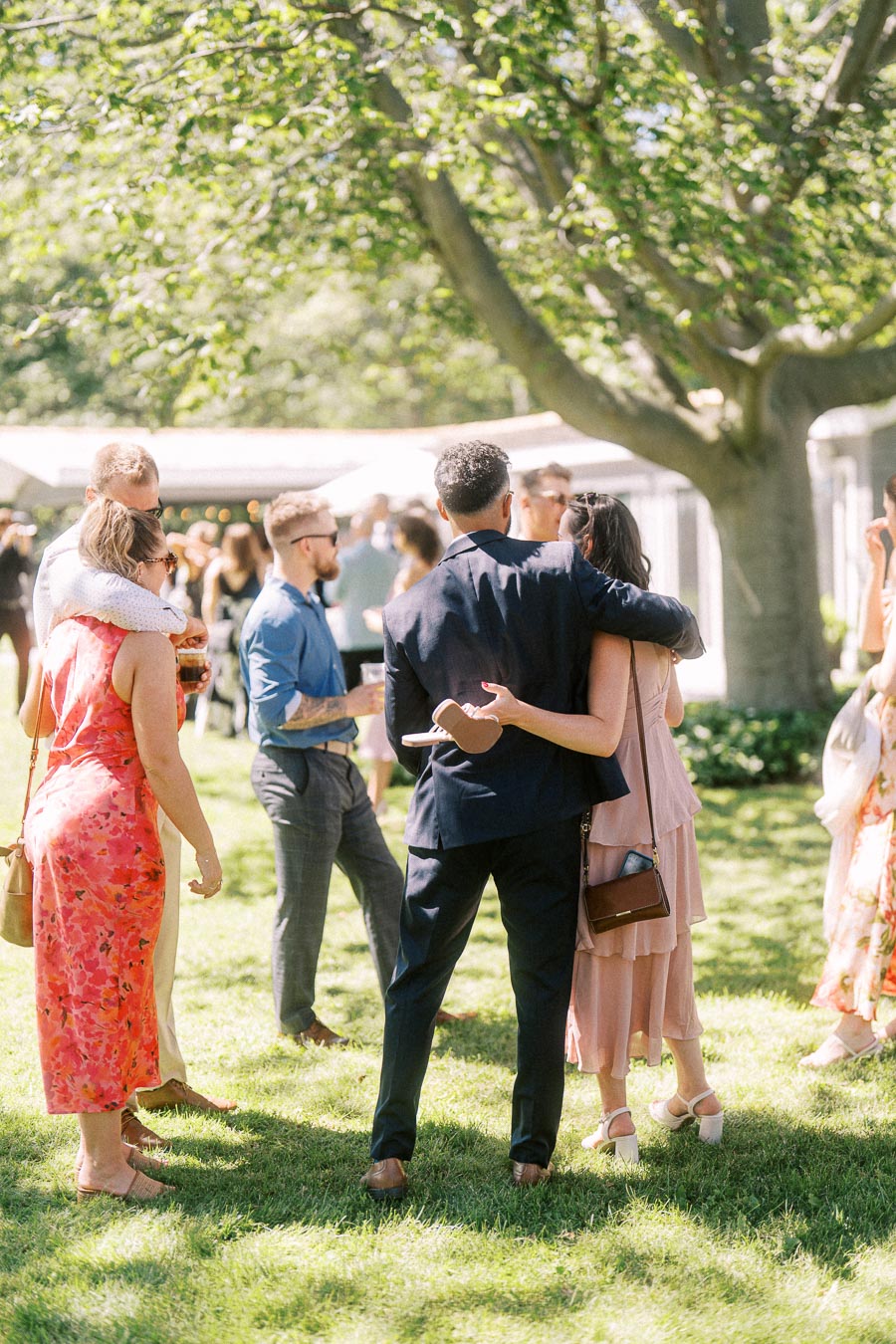 Wedding guests mingle during cocktail hour on a sunny lawn beneath large trees at a private estate outdoor wedding near Chatham-Kent, Ontario, Canada.