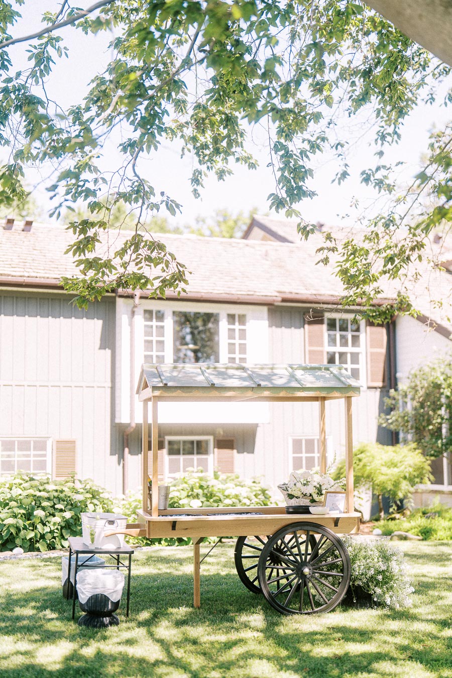 Outdoor wedding cocktail hour drink cart on a private estate lawn near Chatham-Kent, Ontario, with flowers, hydrangeas, and a house in summer light.