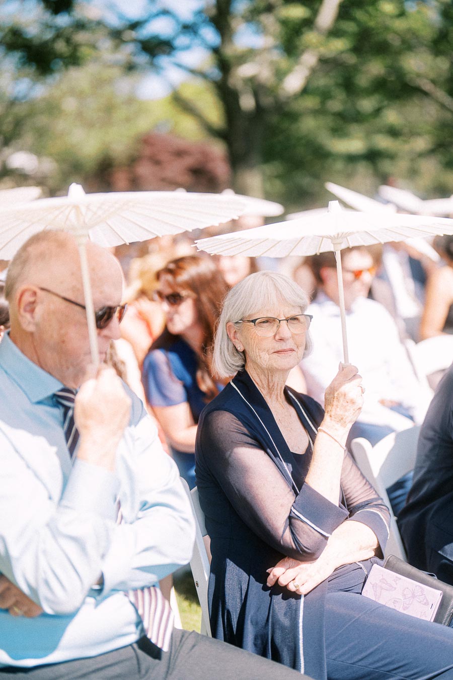 Wedding guests seated outdoors holding white parasols during a sunny private estate ceremony near Chatham-Kent, Ontario, Canada.