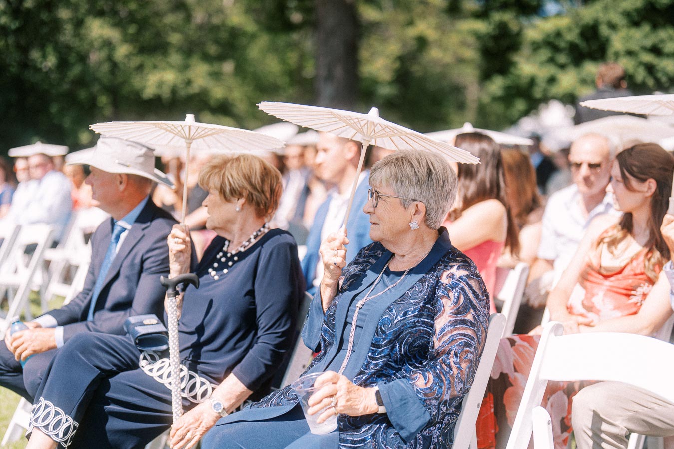 Wedding guests seated outdoors in white chairs holding white parasols during a sunny private estate ceremony near Chatham-Kent, Ontario.