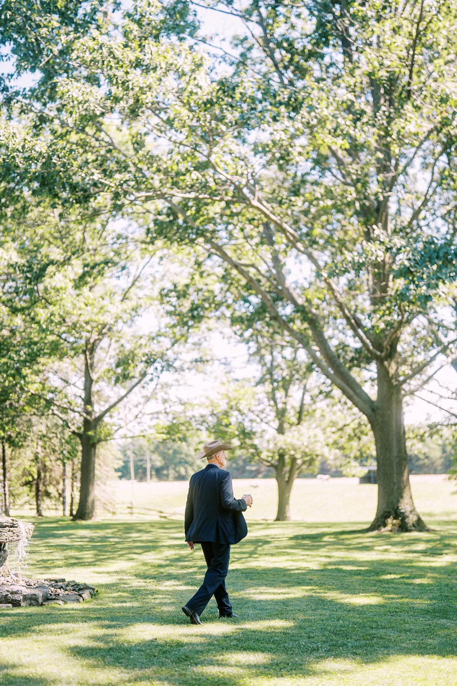 Man in a dark suit and hat walking across a sunlit lawn under large trees at a private estate near Chatham-Kent, Ontario.
