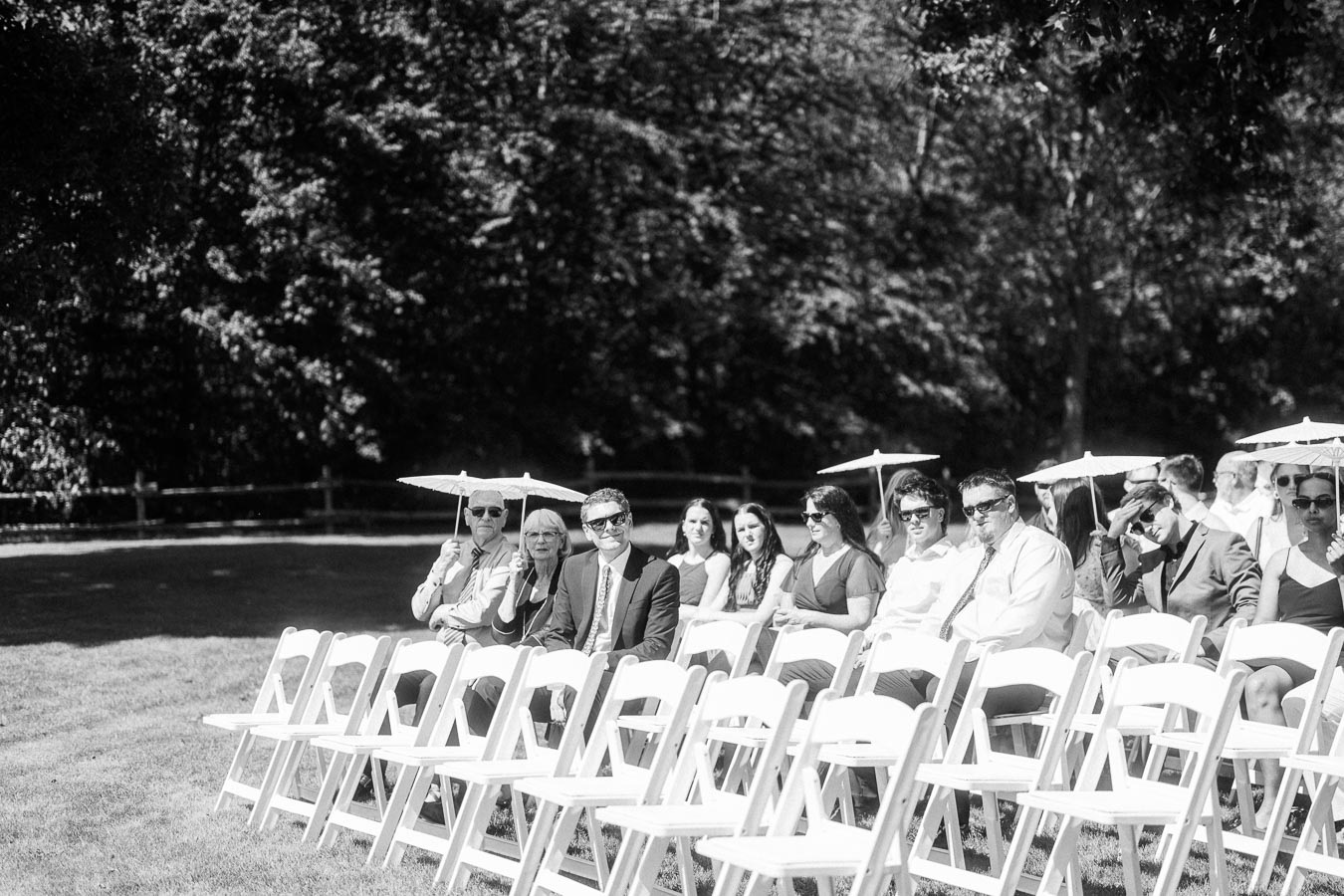 Black and white outdoor wedding ceremony seating with guests on white folding chairs holding parasols on a lawn near Chatham-Kent, Ontario.
