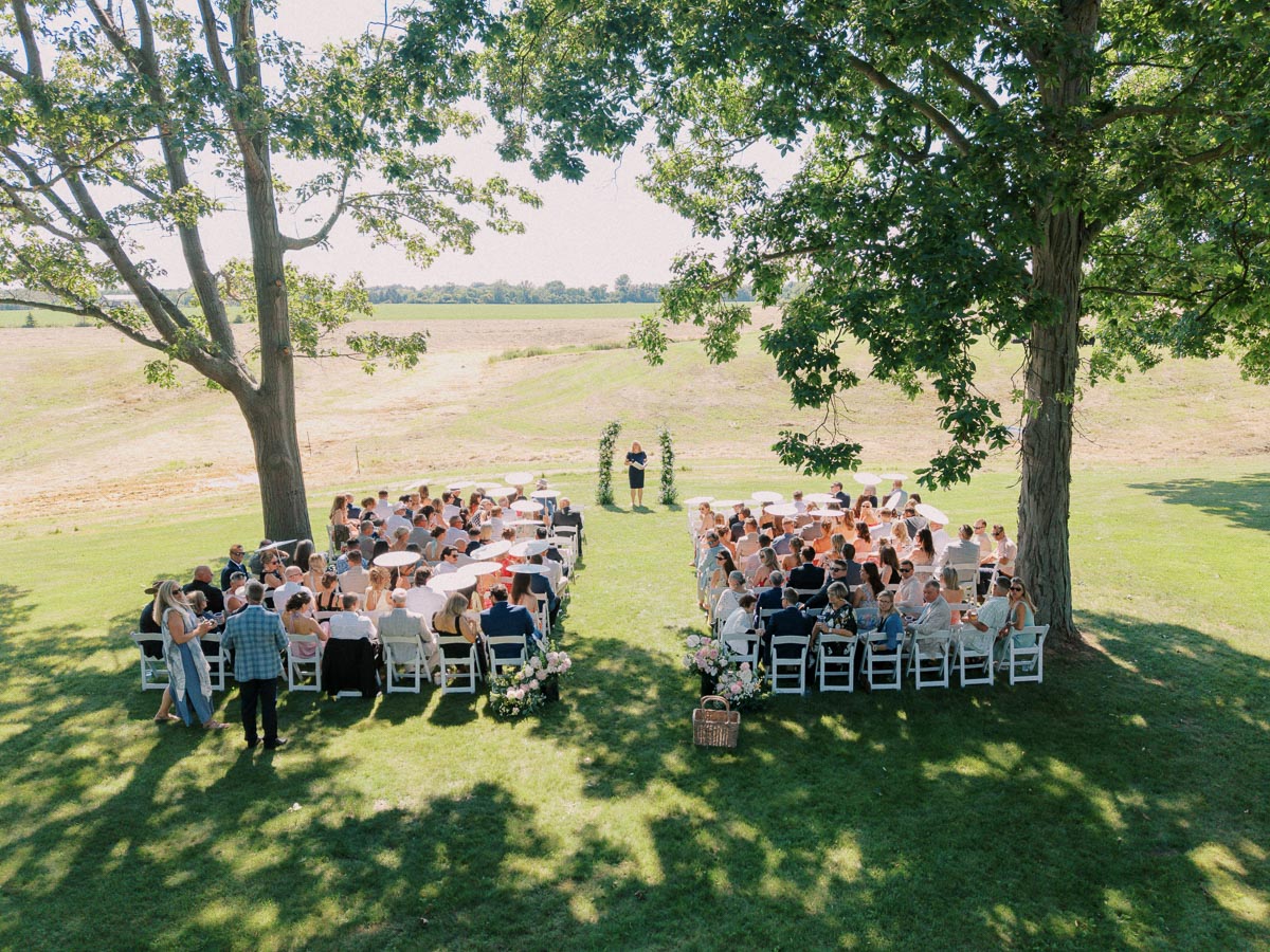 Outdoor wedding ceremony on a private estate lawn near Chatham-Kent, Ontario, with guests seated on white chairs between large trees and farmland.