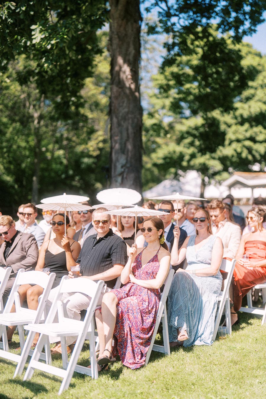 Wedding guests seated on white folding chairs outdoors holding white parasols at a sunny private estate ceremony near Chatham-Kent, Ontario, Canada.