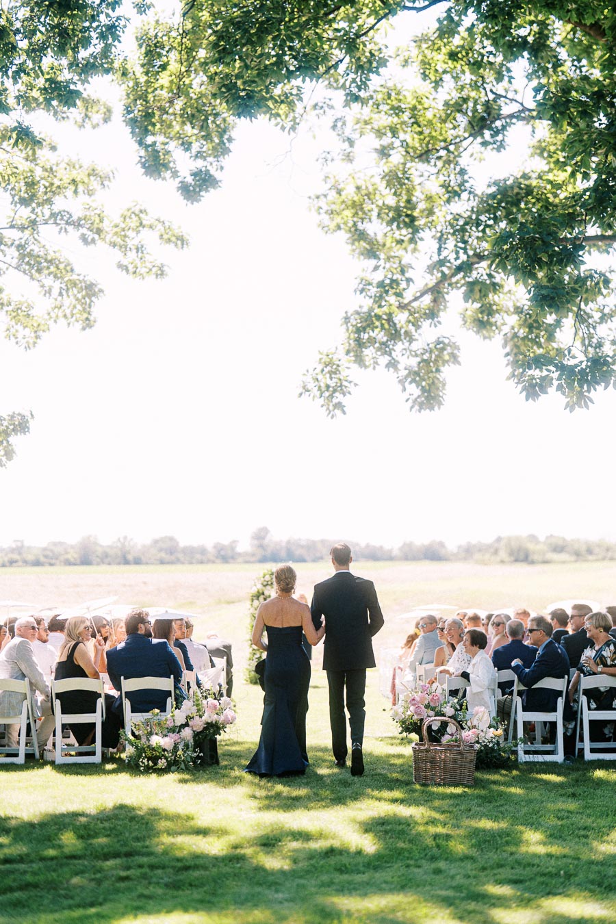 Outdoor wedding ceremony on a private estate near Chatham-Kent, Ontario, with guests seated on white chairs as a couple walks down a floral aisle under trees.