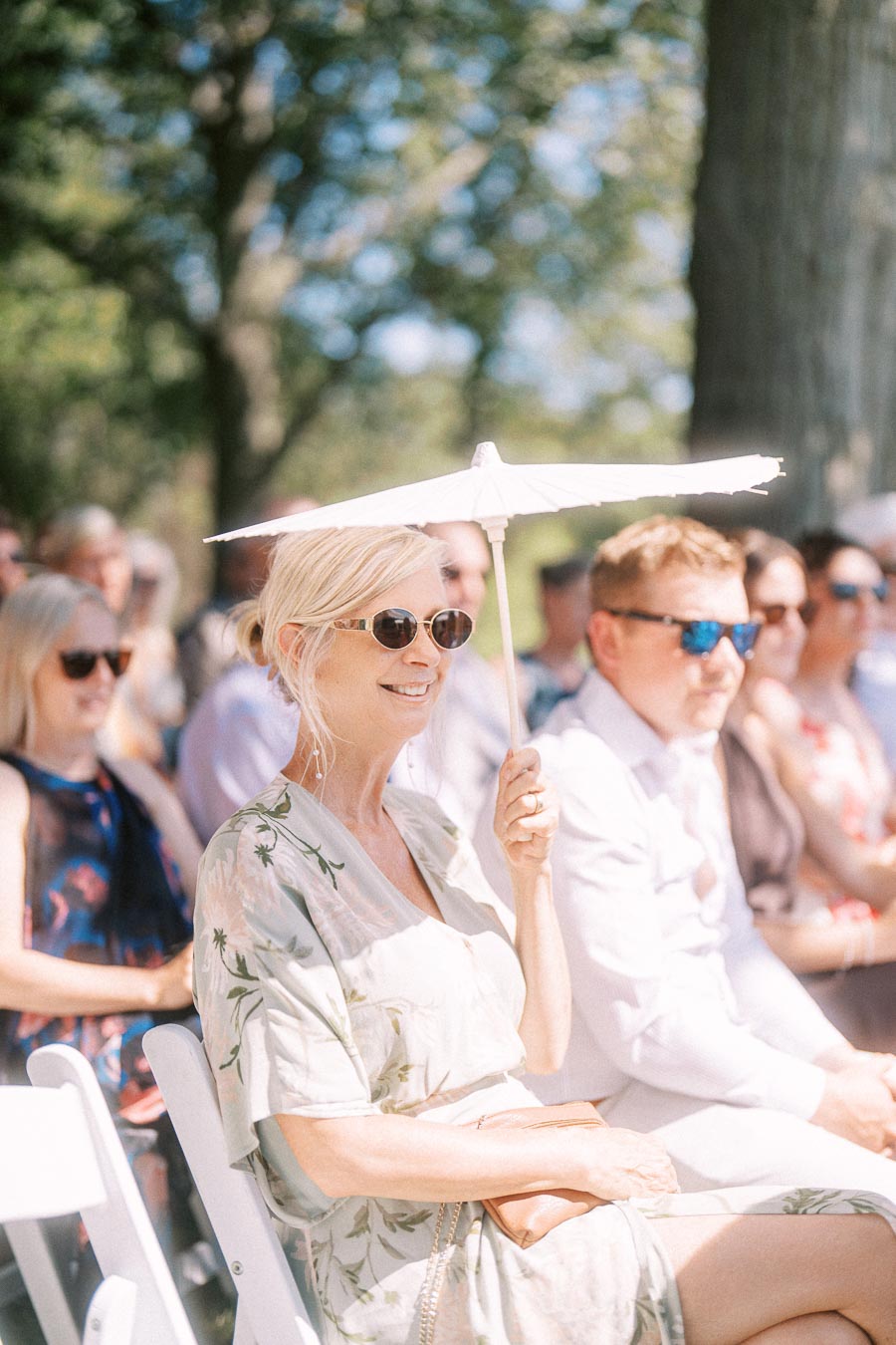 Wedding guests seated outdoors during a sunny ceremony near Chatham-Kent, Ontario, with a woman holding a white parasol for shade.