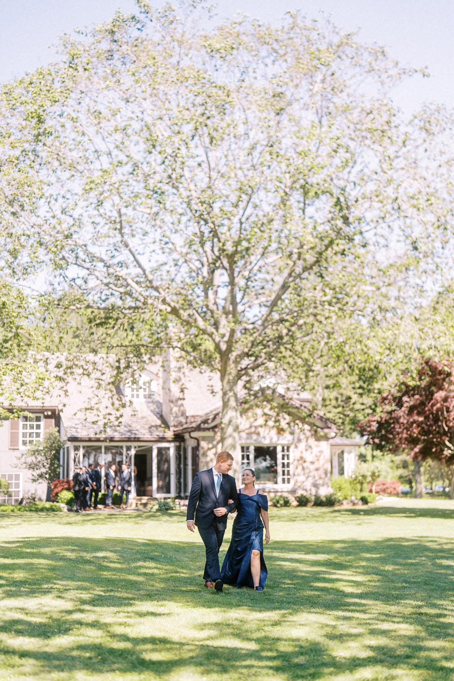 Well dressed wedding guests walk arm in arm across a sunny lawn at a private estate near Chatham-Kent, Ontario, with a house and trees behind them.