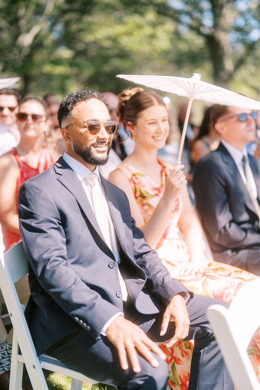 Guests seated outdoors at a sunny wedding ceremony near Chatham-Kent, Ontario, with a man in a suit and sunglasses beside a woman holding a white parasol.