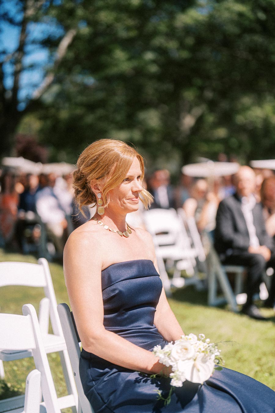 Woman in navy strapless dress holding white flowers at an outdoor wedding ceremony near Chatham-Kent, Ontario, Canada.