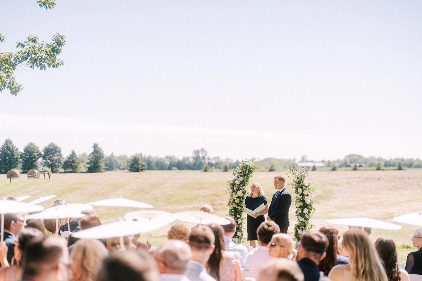 Outdoor wedding ceremony on a private estate near Chatham-Kent, Ontario, with groom, officiant, guests, parasols, floral arch, and open farm field.