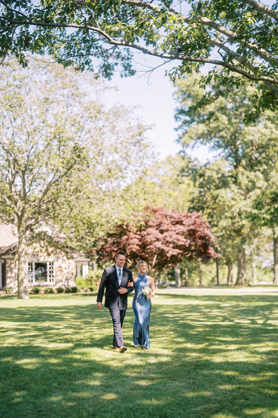 Man in navy suit escorts woman in blue dress holding bouquet across a shaded lawn at an outdoor private estate wedding near Chatham-Kent, Ontario.