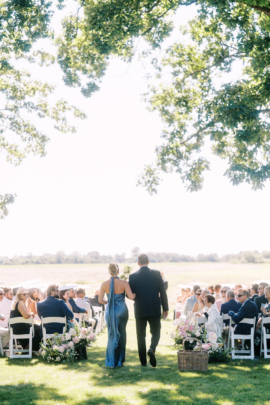Couple walking down a grassy aisle at an outdoor wedding ceremony on a private estate near Chatham-Kent, Ontario, with seated guests, flowers, and trees.
