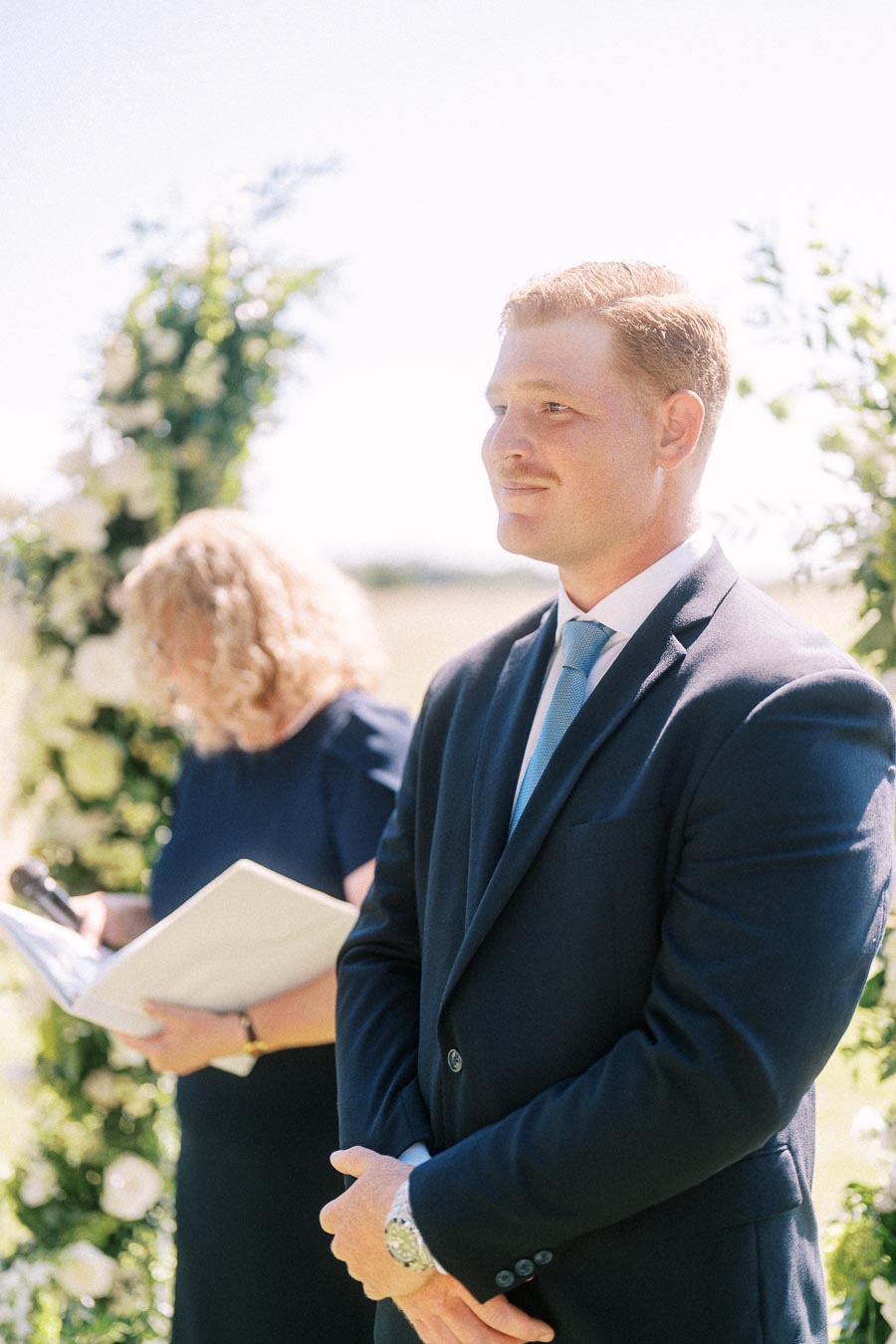 Groom in navy suit standing outdoors during a wedding ceremony near a floral arch while an officiant holds papers in soft sunlight.