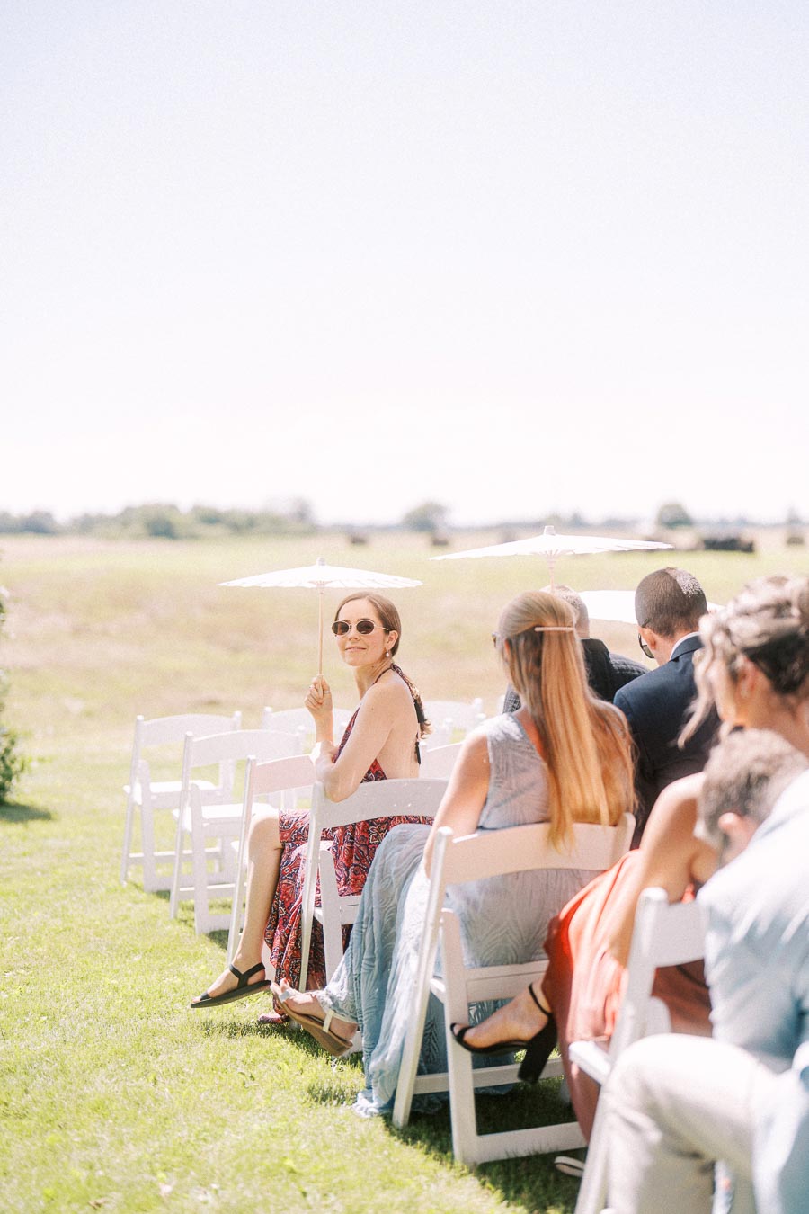 Wedding guests seated on white chairs holding parasols during an outdoor ceremony on a sunny farm field near Chatham-Kent, Ontario, Canada.