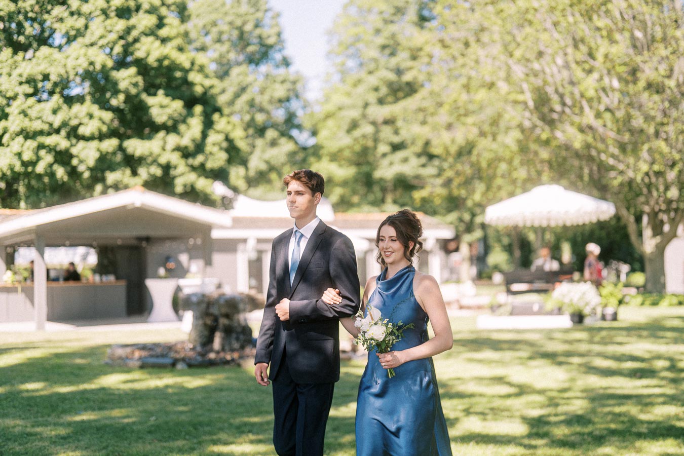 Wedding attendants walking arm in arm on a sunny private estate lawn near Chatham-Kent, Ontario, with bouquet, trees, and tents.