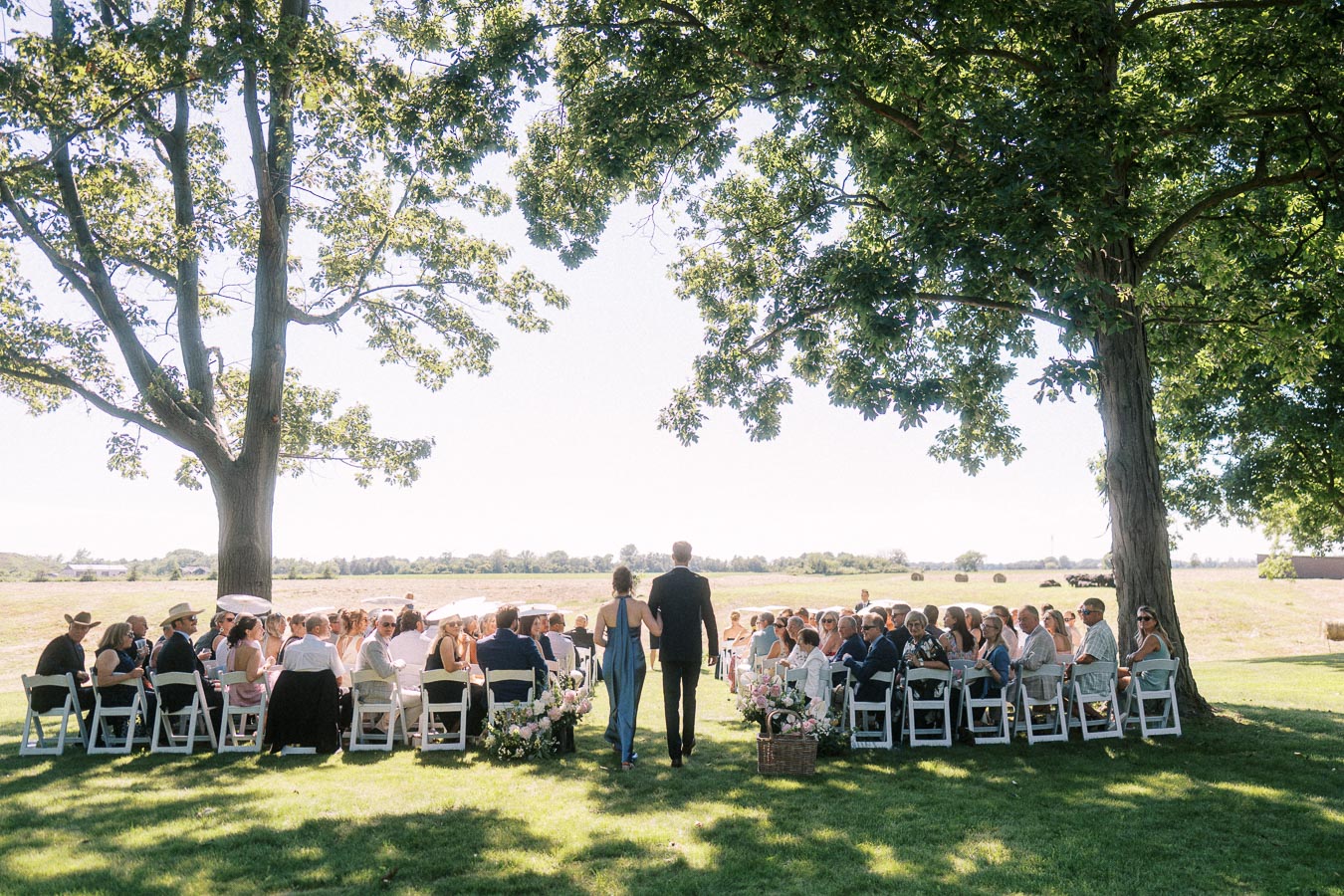 Outdoor private estate wedding ceremony under large trees near Chatham-Kent, Ontario, with guests seated on white chairs beside farmland.