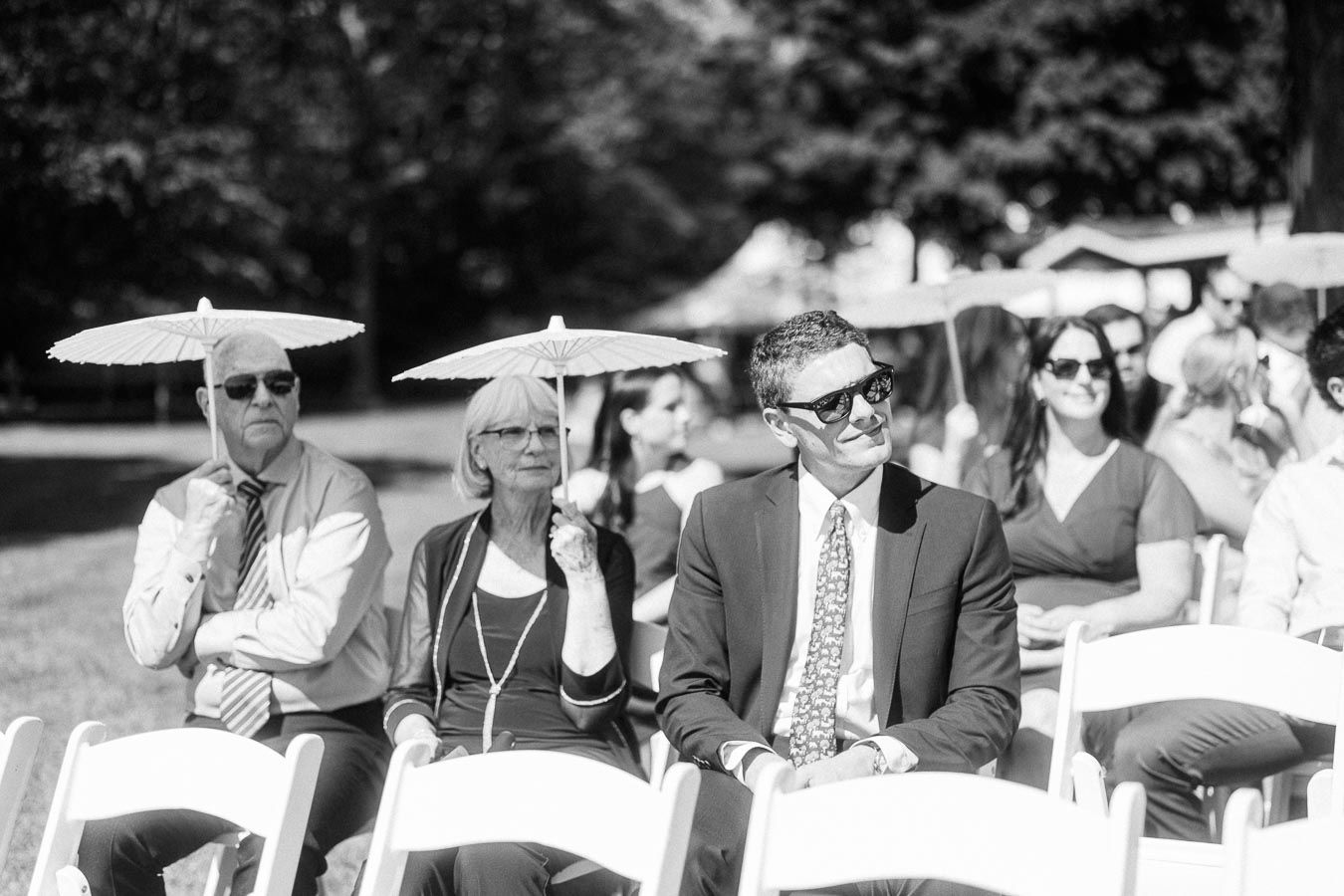 Black and white photo of wedding guests seated on white chairs outdoors, with two guests holding paper parasols for shade near Chatham-Kent, Ontario.