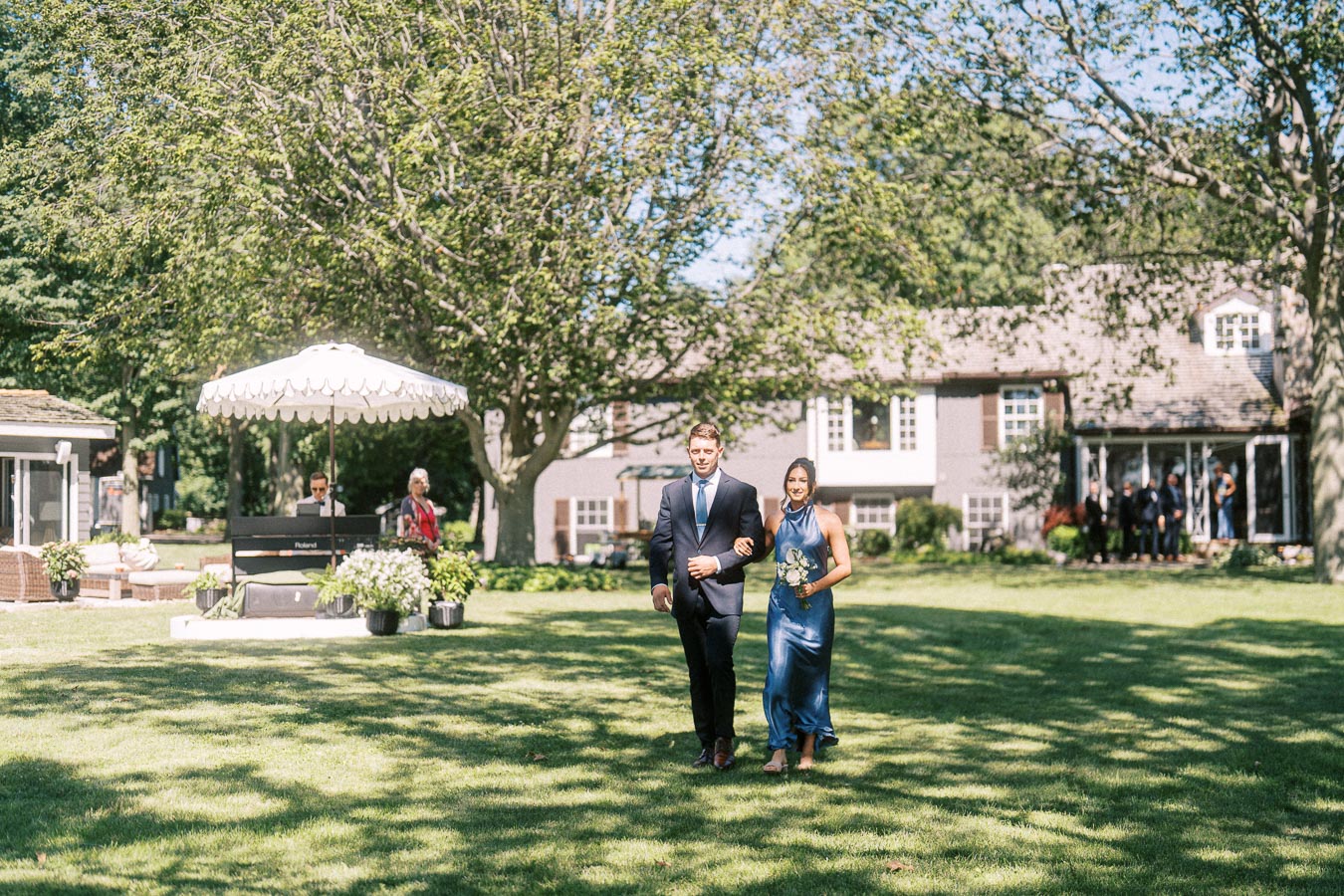 Wedding attendants walk arm in arm across a sunlit lawn during an outdoor private estate ceremony near Chatham-Kent, Ontario.
