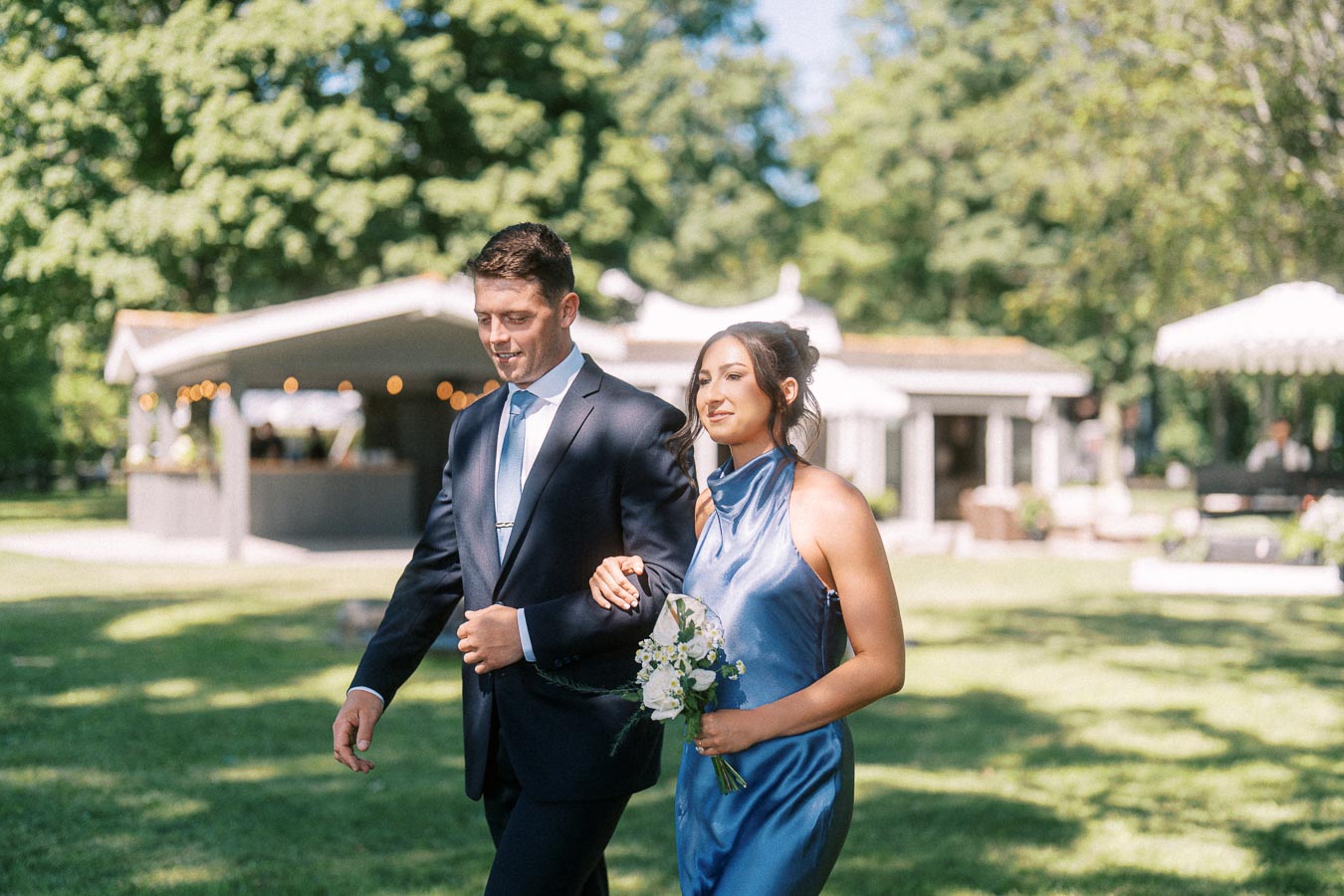 Bridesmaid in blue dress walks arm in arm with groomsman in navy suit at an outdoor private estate wedding in Chatham-Kent, Ontario.