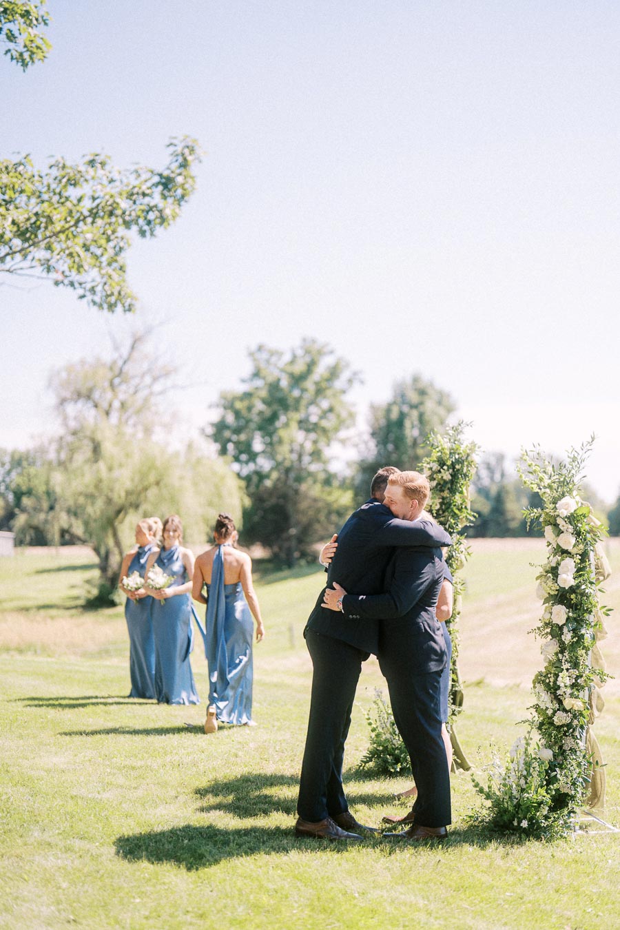 Two men embrace beside a floral wedding arch during an outdoor private estate ceremony near Chatham-Kent, Ontario, with bridesmaids on a lawn.