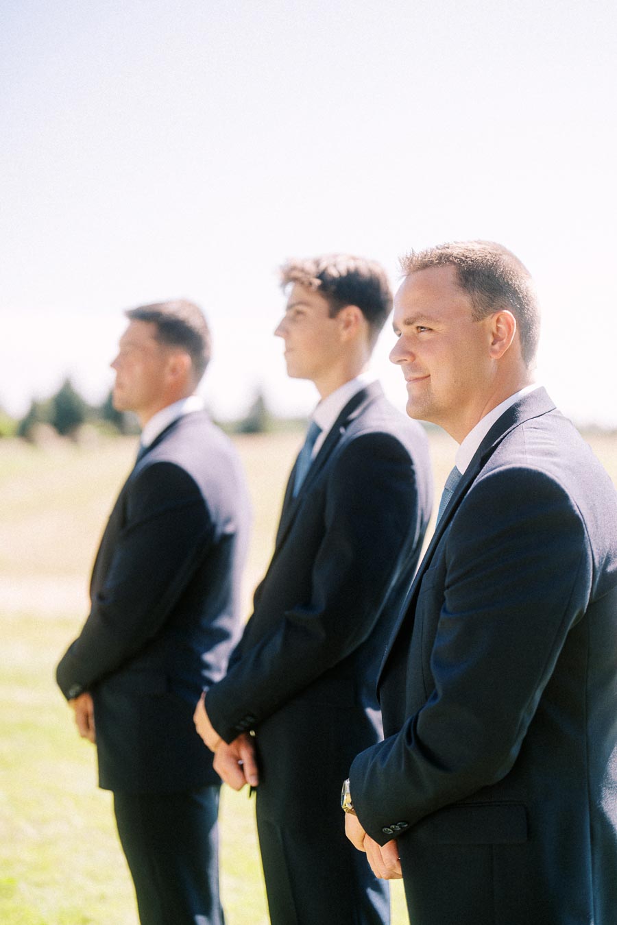Three groomsmen in dark suits standing outdoors in a sunny rural wedding ceremony near Chatham-Kent, Ontario, Canada.