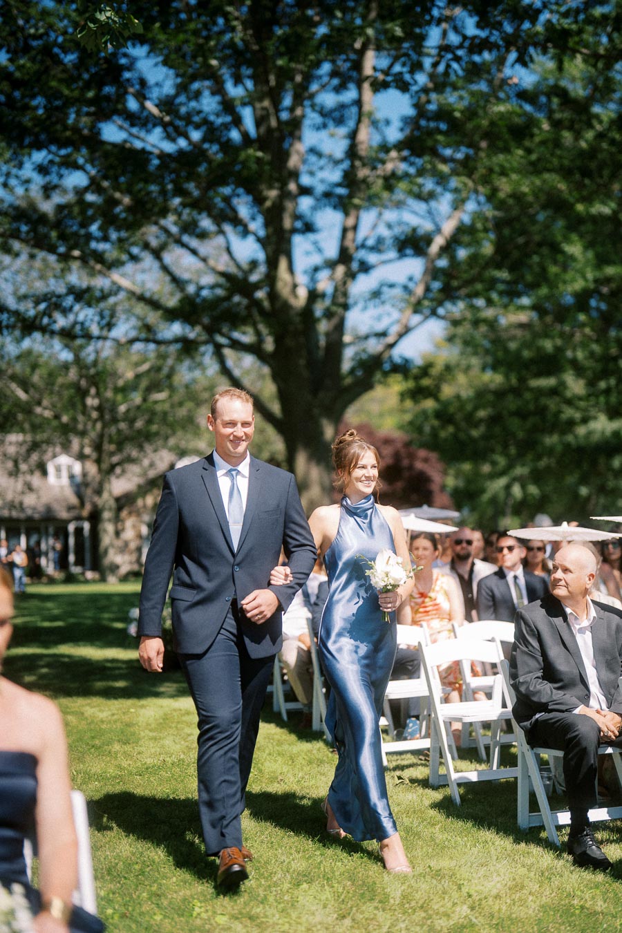 Man in navy suit escorts woman in blue satin dress holding flowers down a grassy aisle at an outdoor wedding ceremony in Chatham-Kent, Ontario.