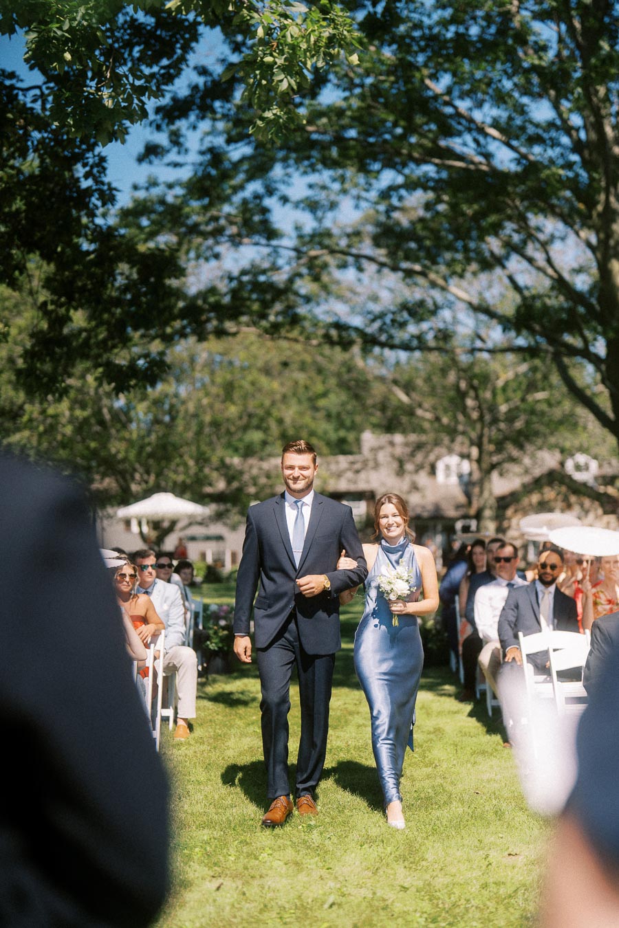 Man in navy suit escorts woman in blue dress with bouquet down a grassy outdoor wedding aisle at a private estate near Chatham-Kent, Ontario.