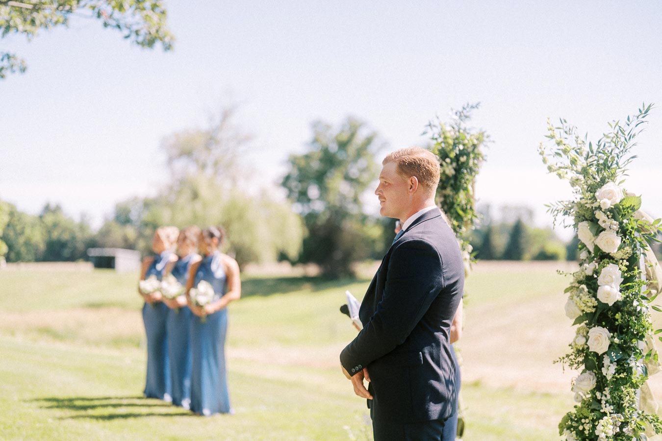 Groom in navy suit stands at an outdoor wedding ceremony beside a white floral arch on a private estate near Chatham-Kent, Ontario.