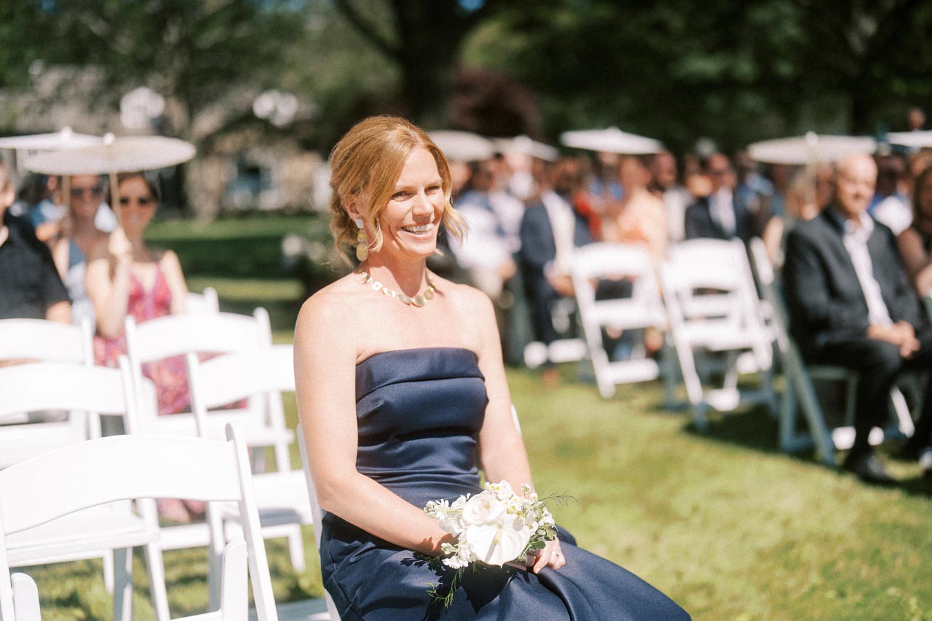 Smiling red haired woman in a navy dress holding a white bouquet while seated among guests at an outdoor wedding ceremony in Chatham-Kent, Ontario.
