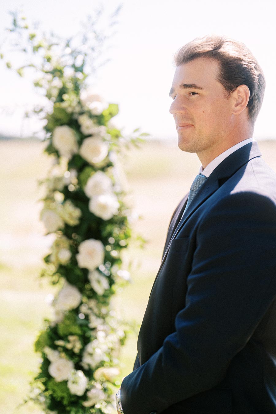 Groom in a navy suit standing at an outdoor wedding ceremony beside a white floral arch in Chatham-Kent, Ontario.