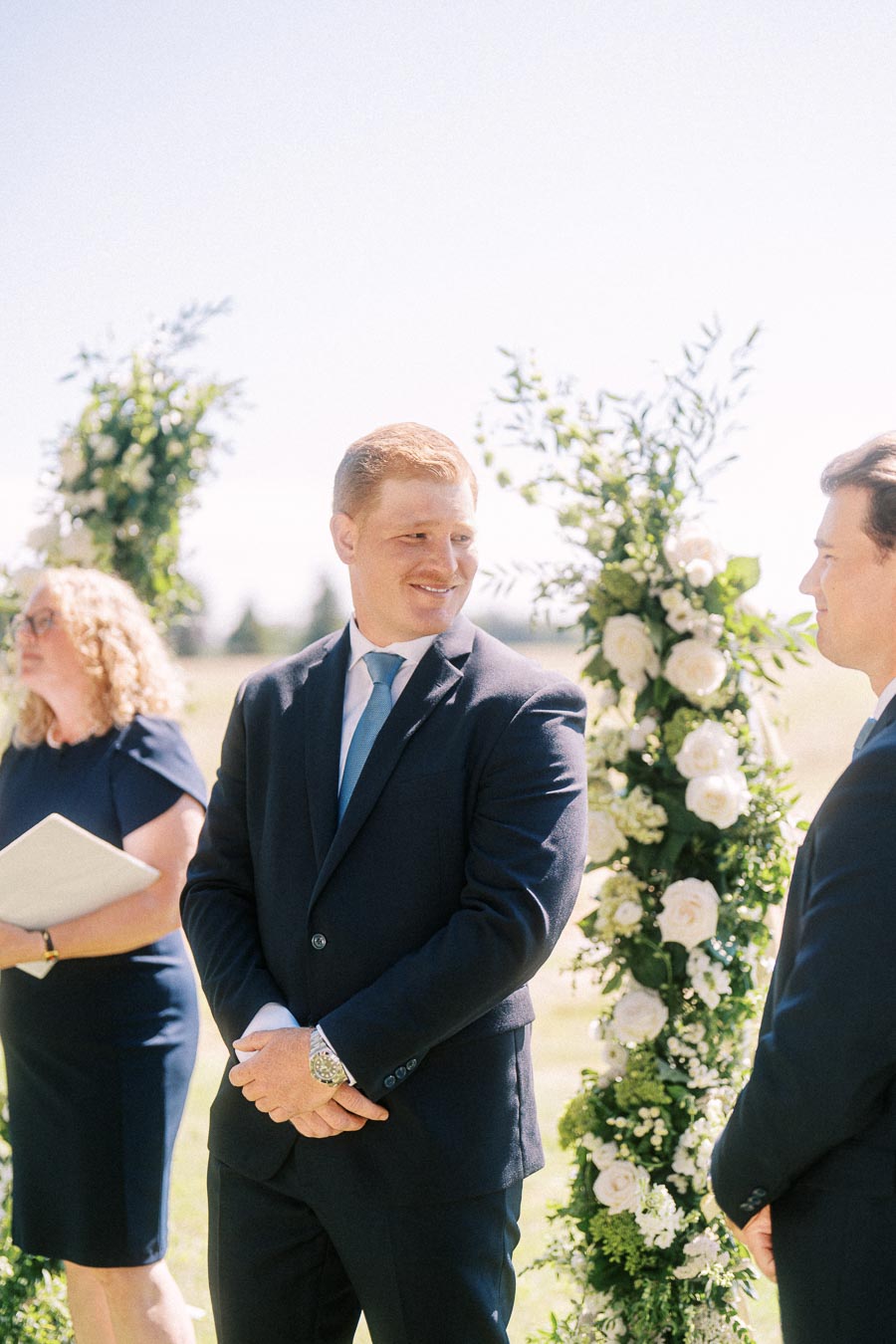 Groom in a navy suit smiling during an outdoor wedding ceremony beside a white floral arch in Chatham-Kent, Ontario, Canada.