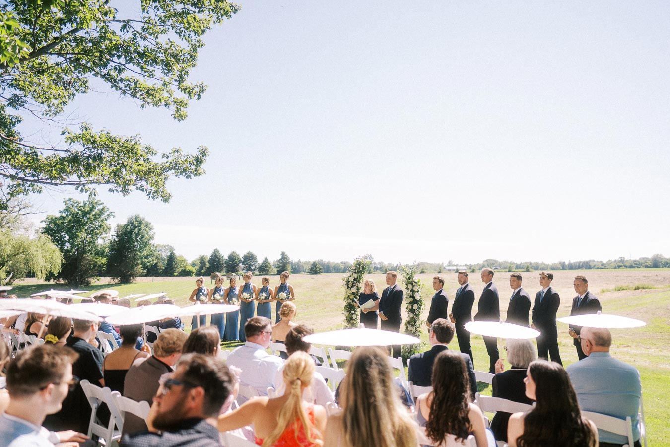 Outdoor wedding ceremony on a private estate near Chatham-Kent, Ontario, with guests seated in white chairs facing the wedding party in a sunny field.