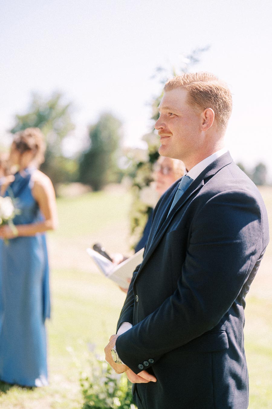Groom in a dark suit smiling during an outdoor wedding ceremony on a sunny private estate near Chatham-Kent, Ontario, Canada.