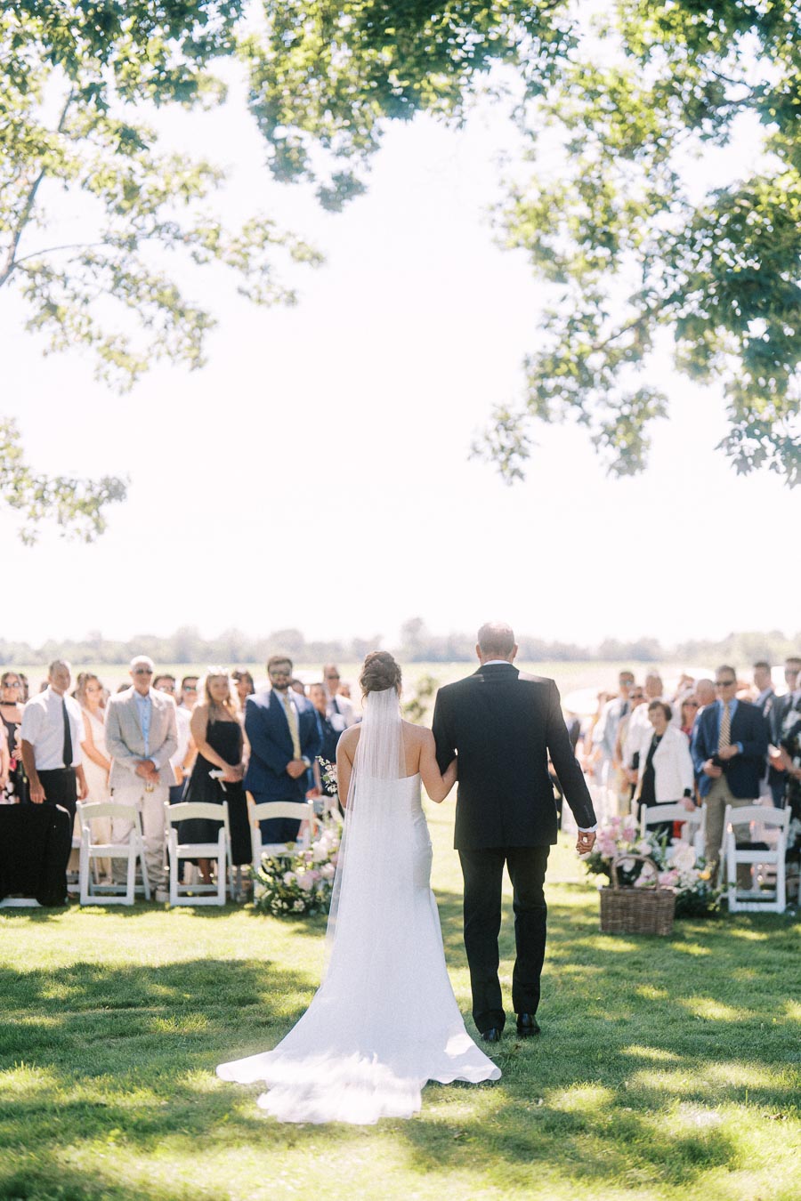 Bride in white gown walks down an outdoor wedding aisle with an older man as guests stand beside chairs on a private estate lawn near Chatham-Kent, Ontario.
