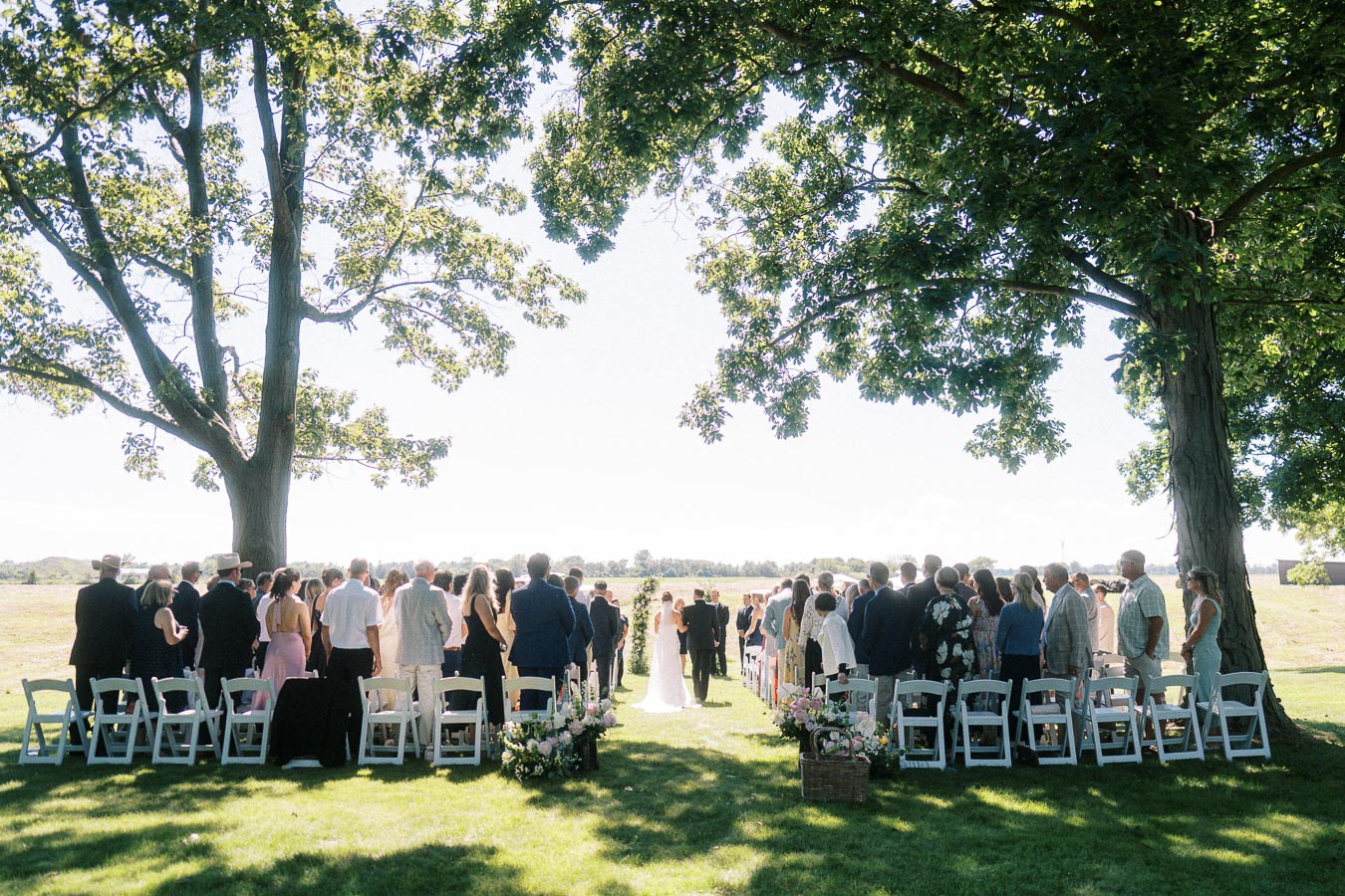 Outdoor wedding ceremony with guests standing under large trees on a private estate near Chatham-Kent, Ontario, Canada.