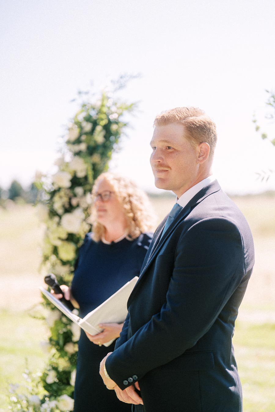 Groom in a navy suit stands beside an officiant at a sunny outdoor wedding ceremony near Chatham-Kent, Ontario, Canada.