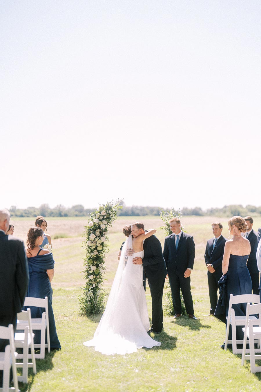 Bride and groom kiss during an outdoor wedding ceremony on a grassy private estate near Chatham-Kent, Ontario, with guests and a floral arch.