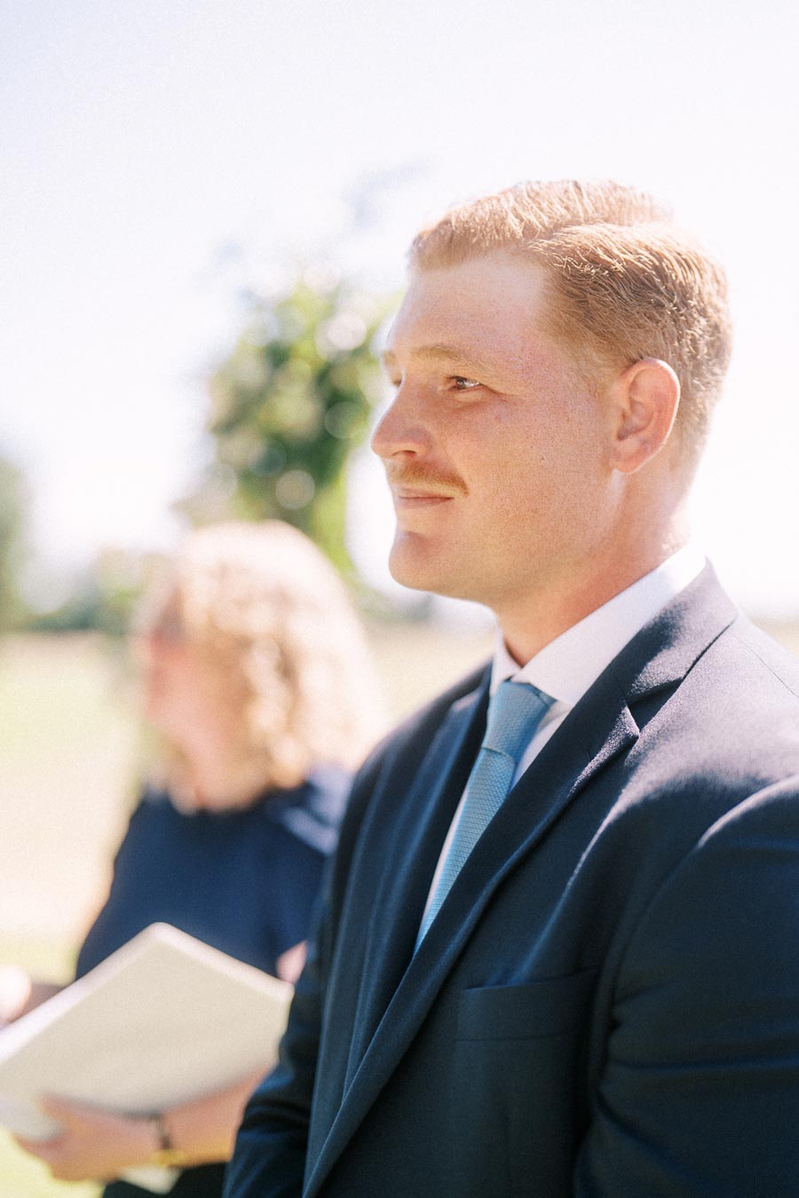 Groom in a navy suit standing outdoors during a private estate wedding ceremony near Chatham-Kent, Ontario, Canada, with an officiant blurred behind him.