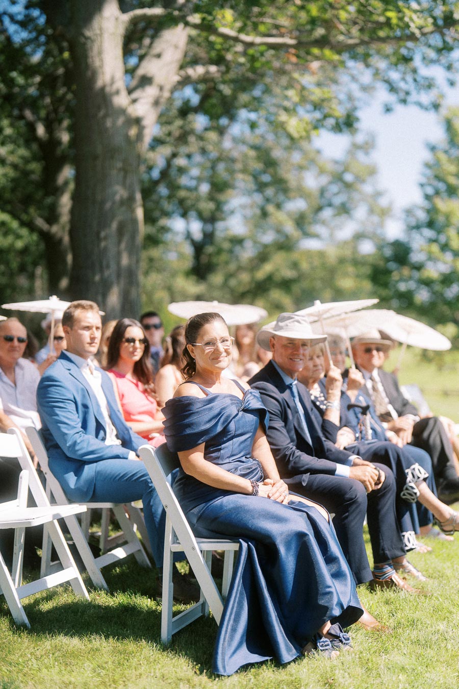 Wedding guests seated on white chairs outdoors at a private estate ceremony near Chatham-Kent, Ontario, with parasols and trees.