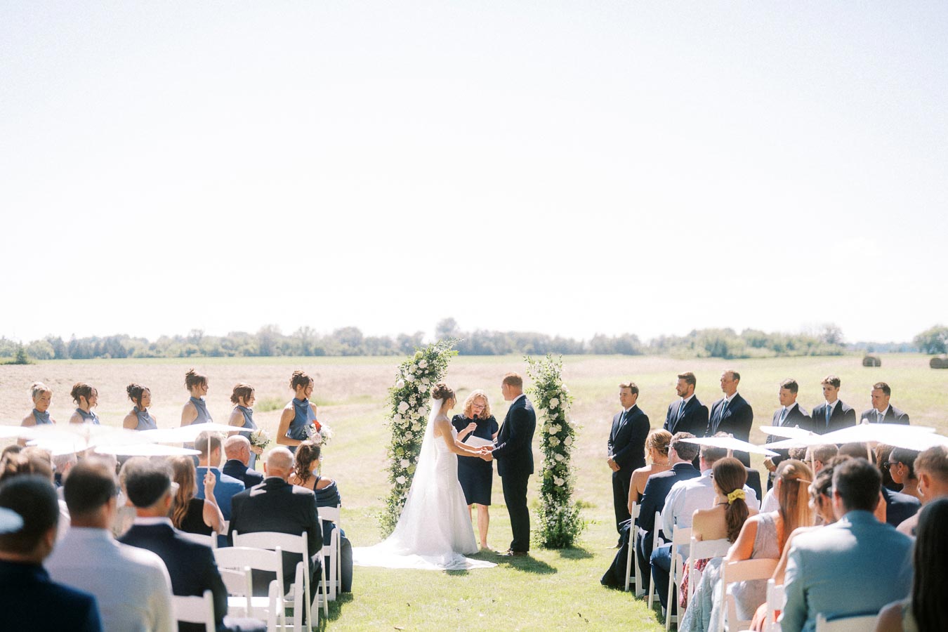 Outdoor private estate wedding ceremony near Chatham-Kent, Ontario, with bride and groom at a floral arch, seated guests, bridesmaids, and groomsmen in a farm field setting.