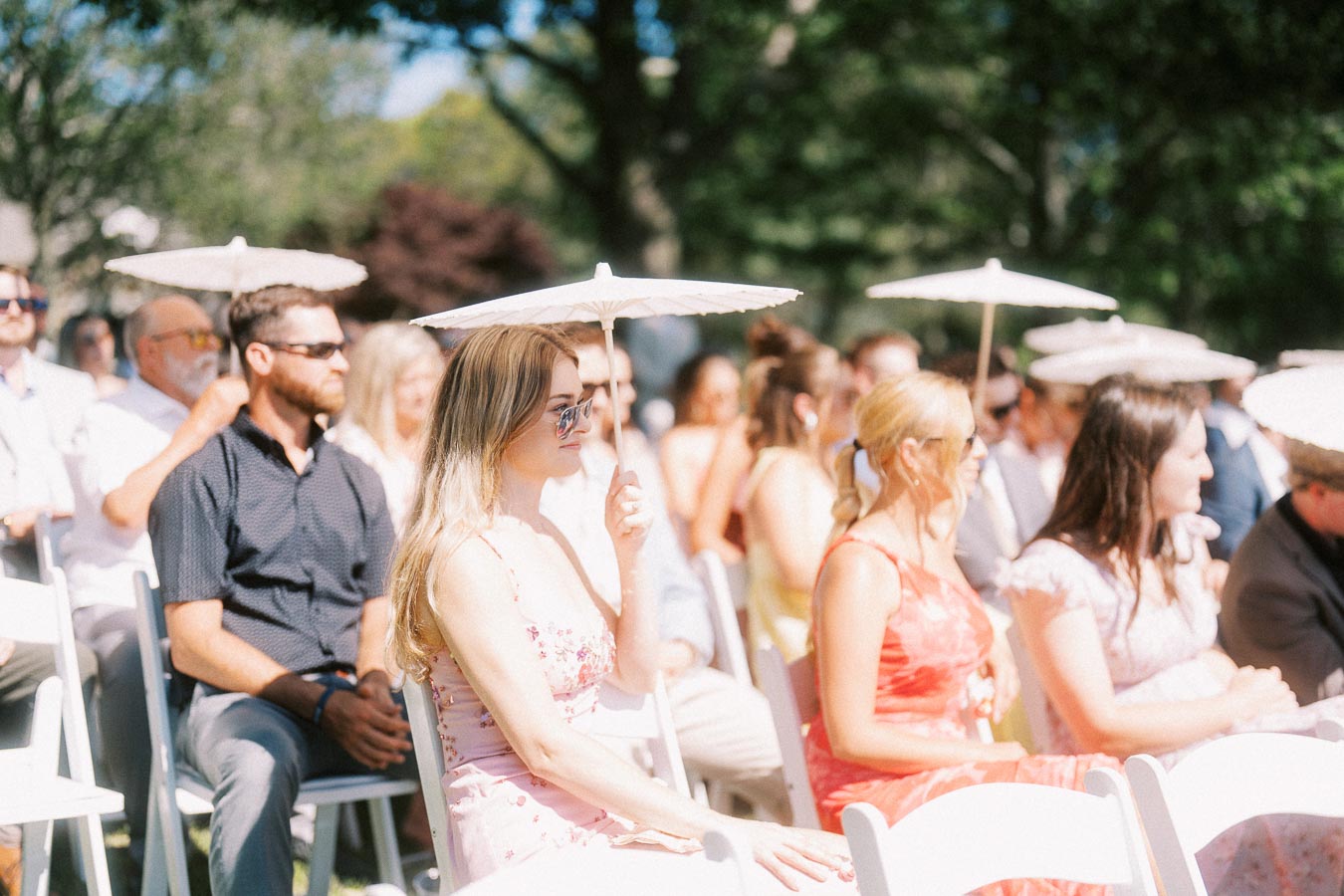 Wedding guests seated outdoors on white chairs holding white parasols during a sunny private estate ceremony near Chatham-Kent, Ontario, Canada.