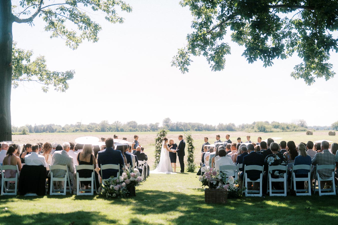 Outdoor wedding ceremony on a private estate lawn near Chatham-Kent, Ontario, with guests seated under trees facing a floral arch and open farm fields.
