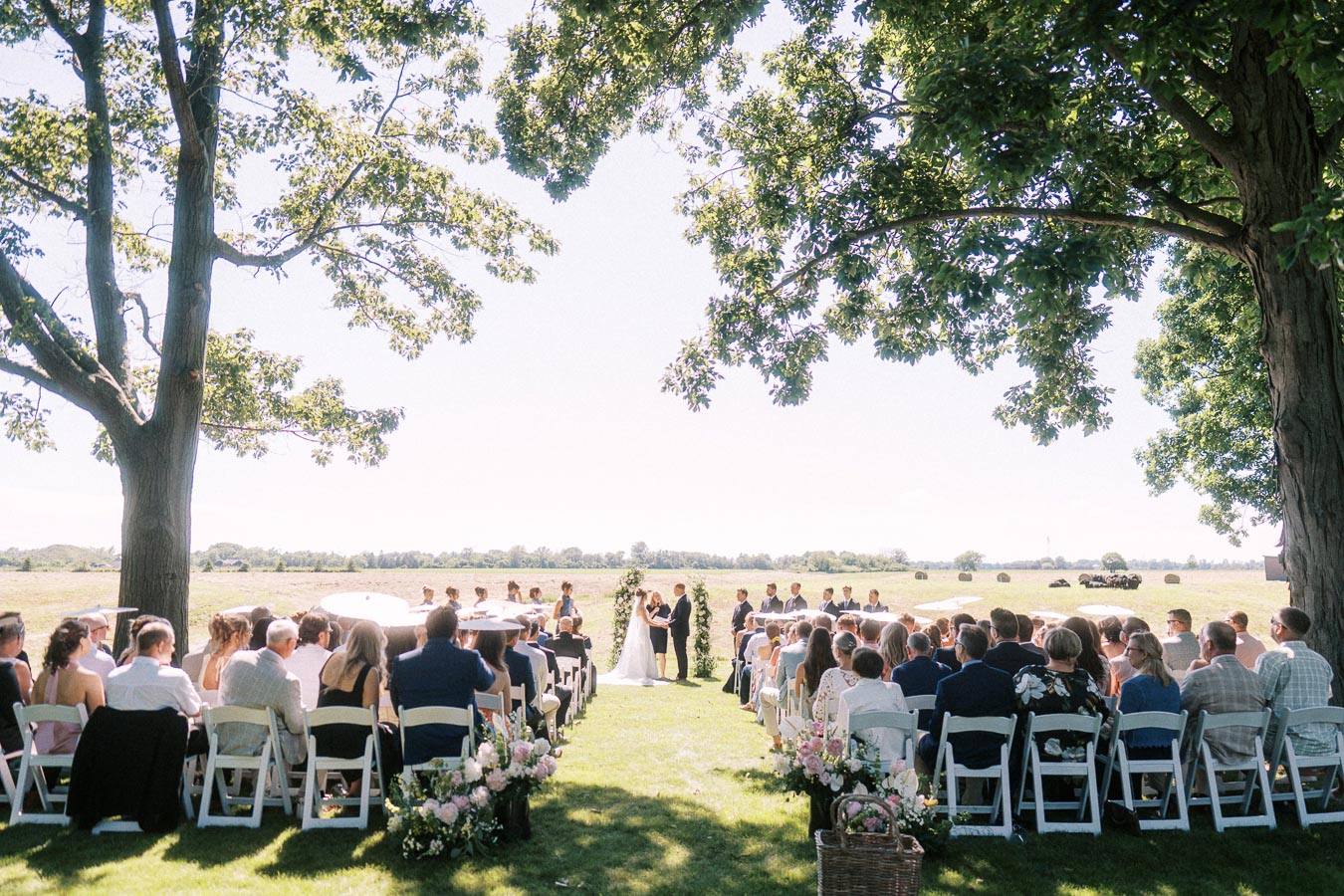 Outdoor wedding ceremony on a private estate near Chatham-Kent, Ontario, with guests seated under large trees beside open farmland.