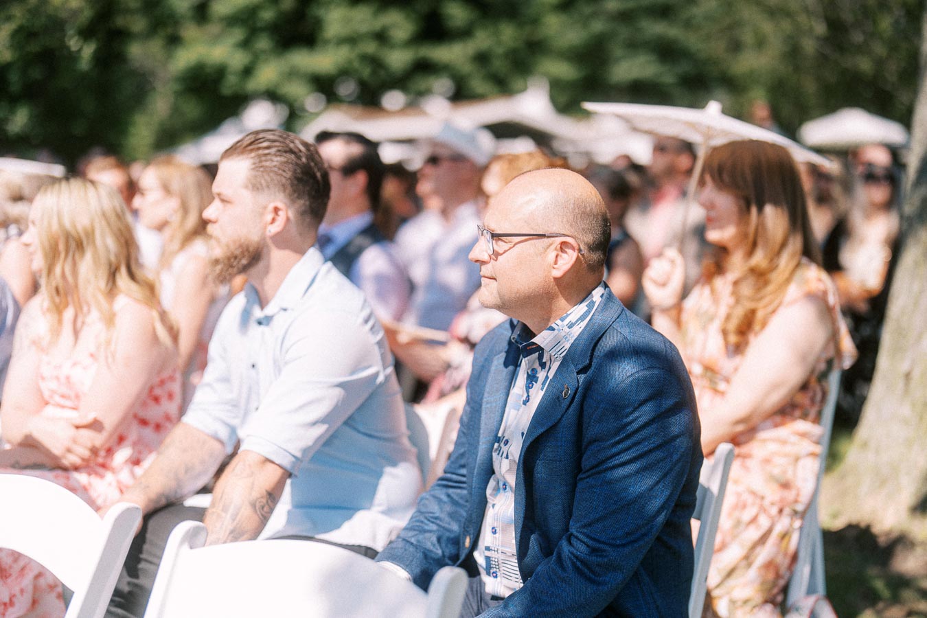 Wedding guests seated on white chairs during an outdoor ceremony near Chatham-Kent, Ontario, with trees and umbrellas in bright sunlight.
