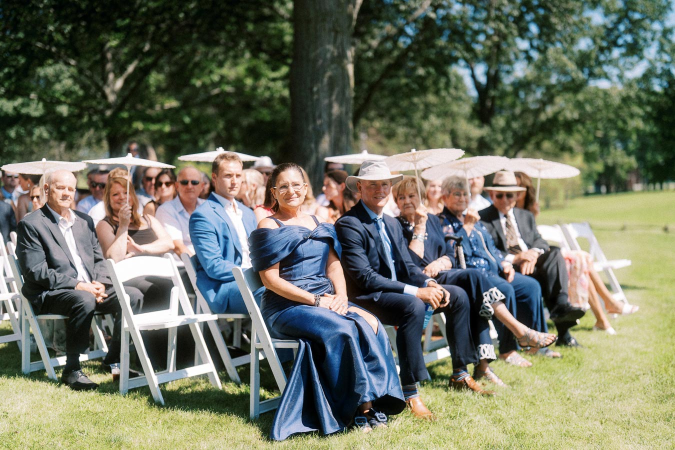 Wedding guests seated on white chairs with parasols during an outdoor ceremony on a private estate lawn near Chatham-Kent, Ontario, Canada.