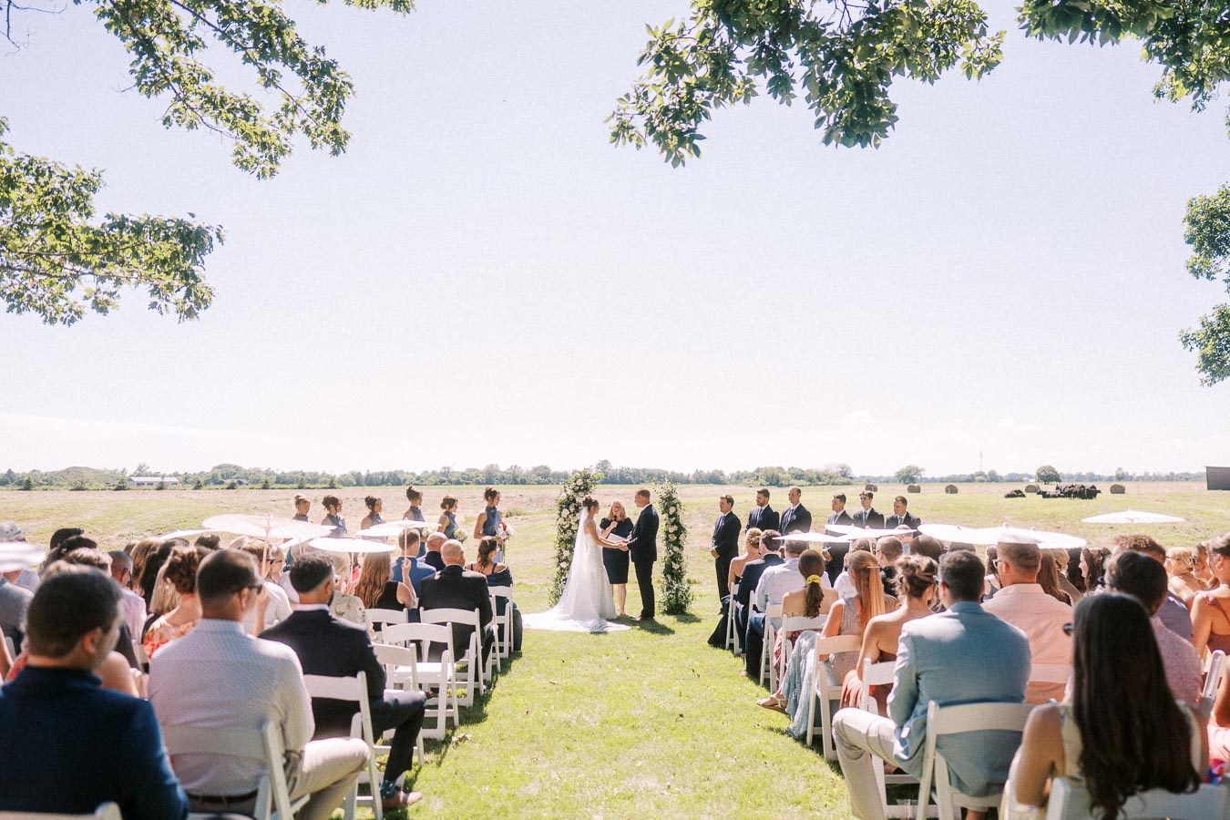 Outdoor wedding ceremony on a private farm estate near Chatham-Kent, Ontario, with guests seated on white chairs under a sunny sky.