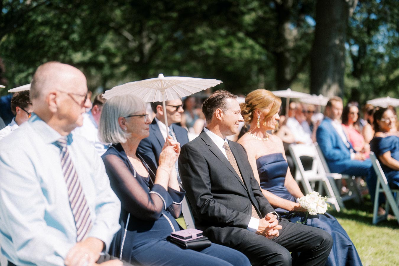 Wedding guests seated outdoors on white chairs during a sunny ceremony near Chatham-Kent, Ontario, with trees and parasols in the background.